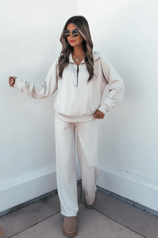 Woman wearing sunglasses, a light beige zip-up, Stone Soft Cloud Scuba Sweatpants, and beige shoes against a white wall.
