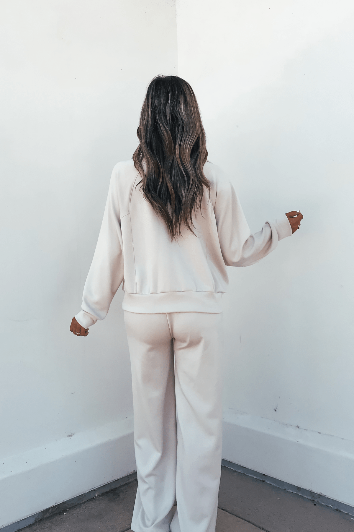 A woman with wavy hair faces a white wall, wearing Stone Soft Cloud Scuba Sweatpants in relaxed fit and light beige.