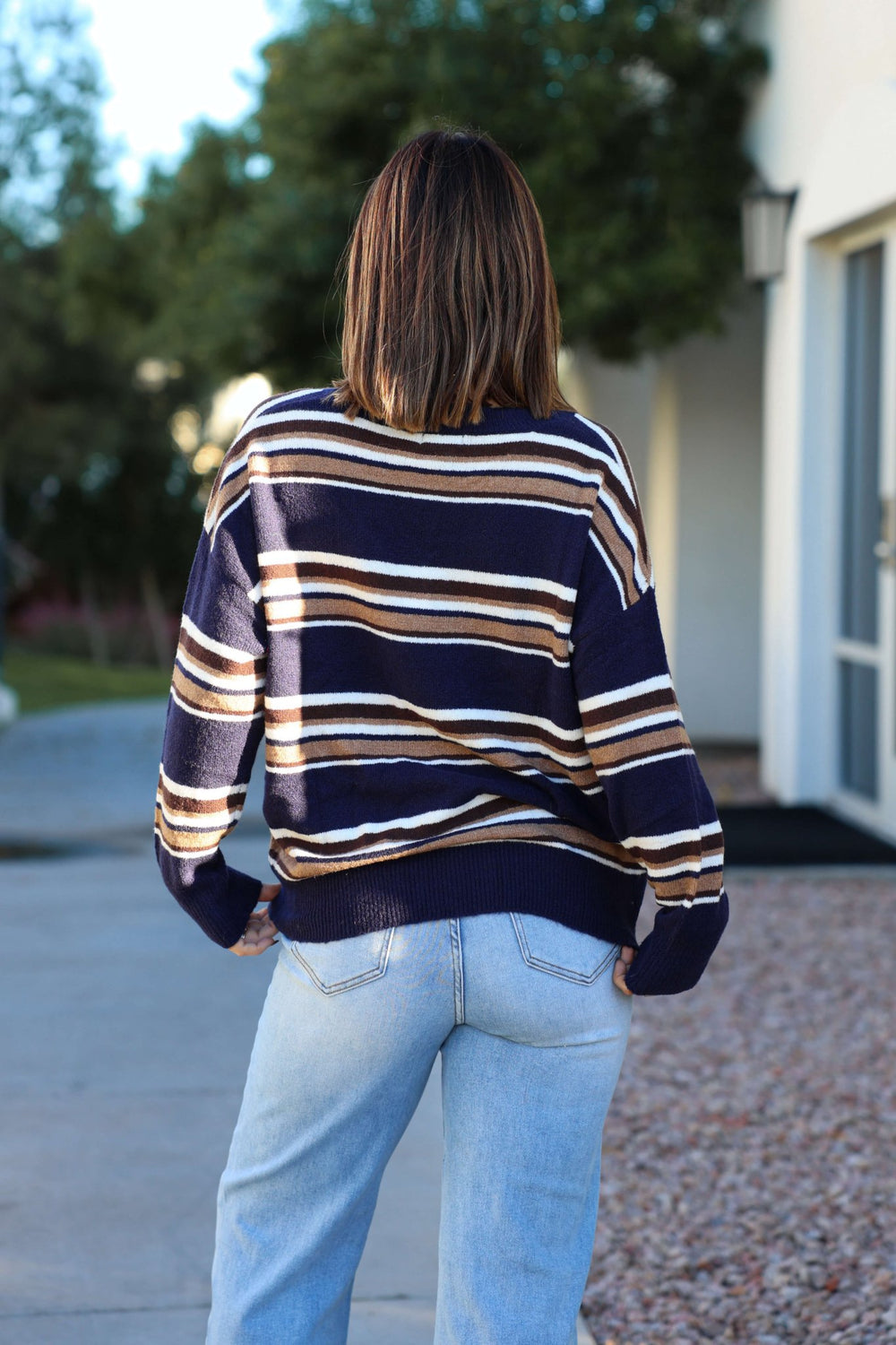 A person with shoulder-length hair wears the Street Chic Multi Navy Striped Sweater and light jeans, standing outside facing away.