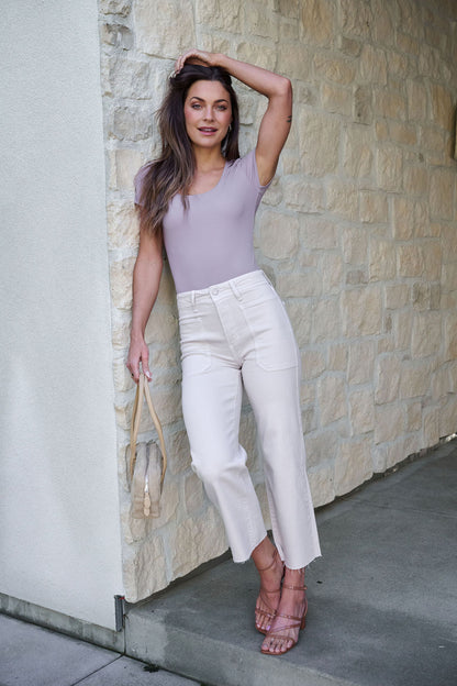 Woman with long hair in a grey Stretchy Basic Scoop Neck Bodysuit and white pants leans against a stone wall, holding a beige bag.