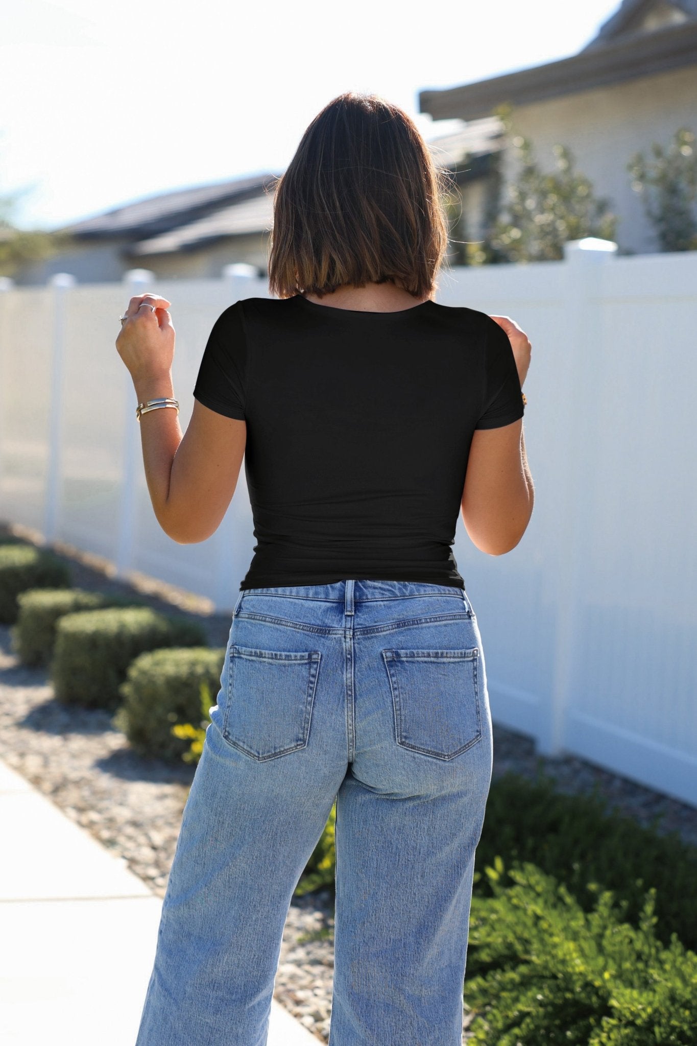 A woman in a Stretchy Black Double Lined Scoop Neck Top and blue jeans stands outside near a white fence, facing away.