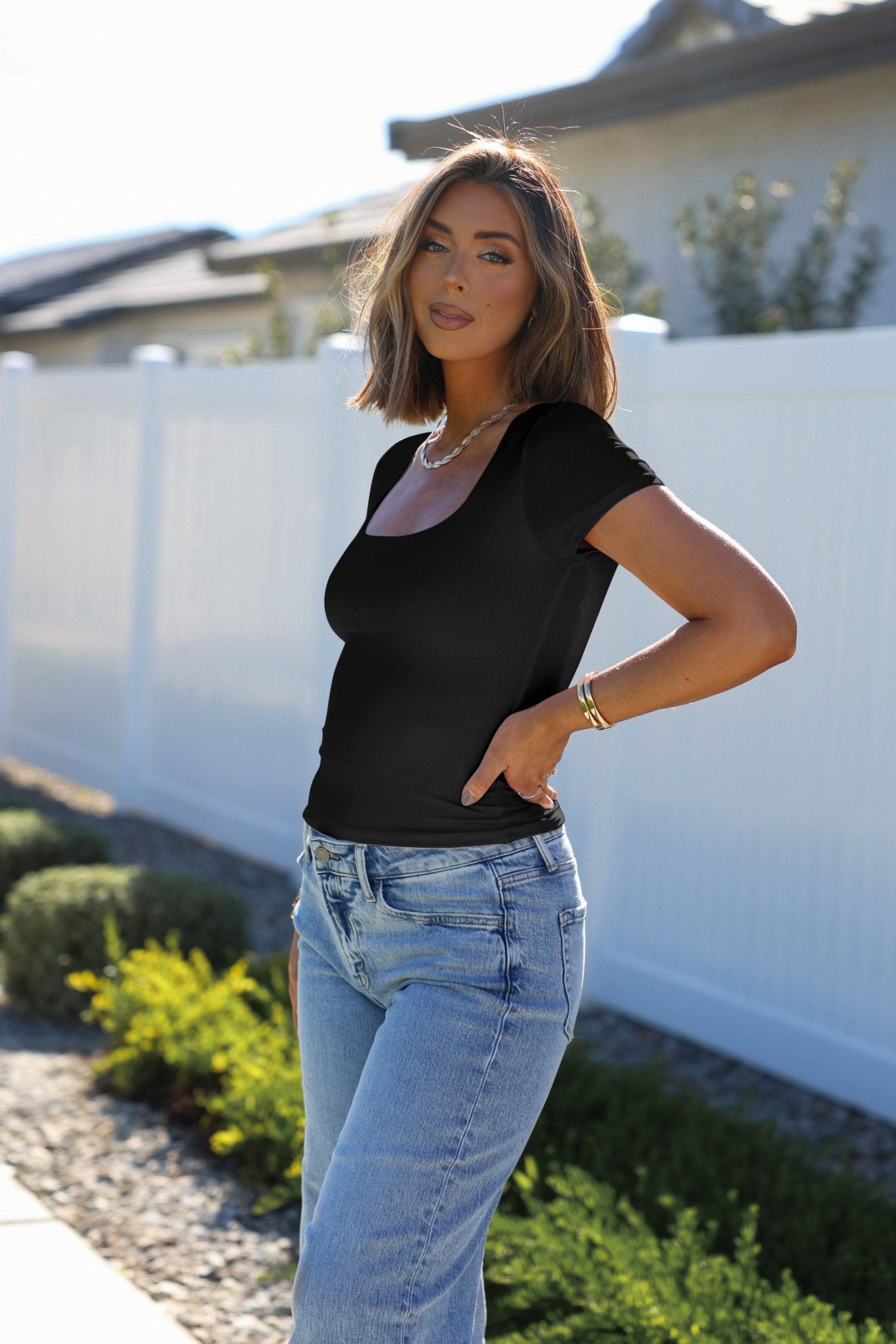 Woman with shoulder-length brown hair wears a Stretchy Black Double Lined Scoop Neck Top and blue jeans, posing by a white fence.