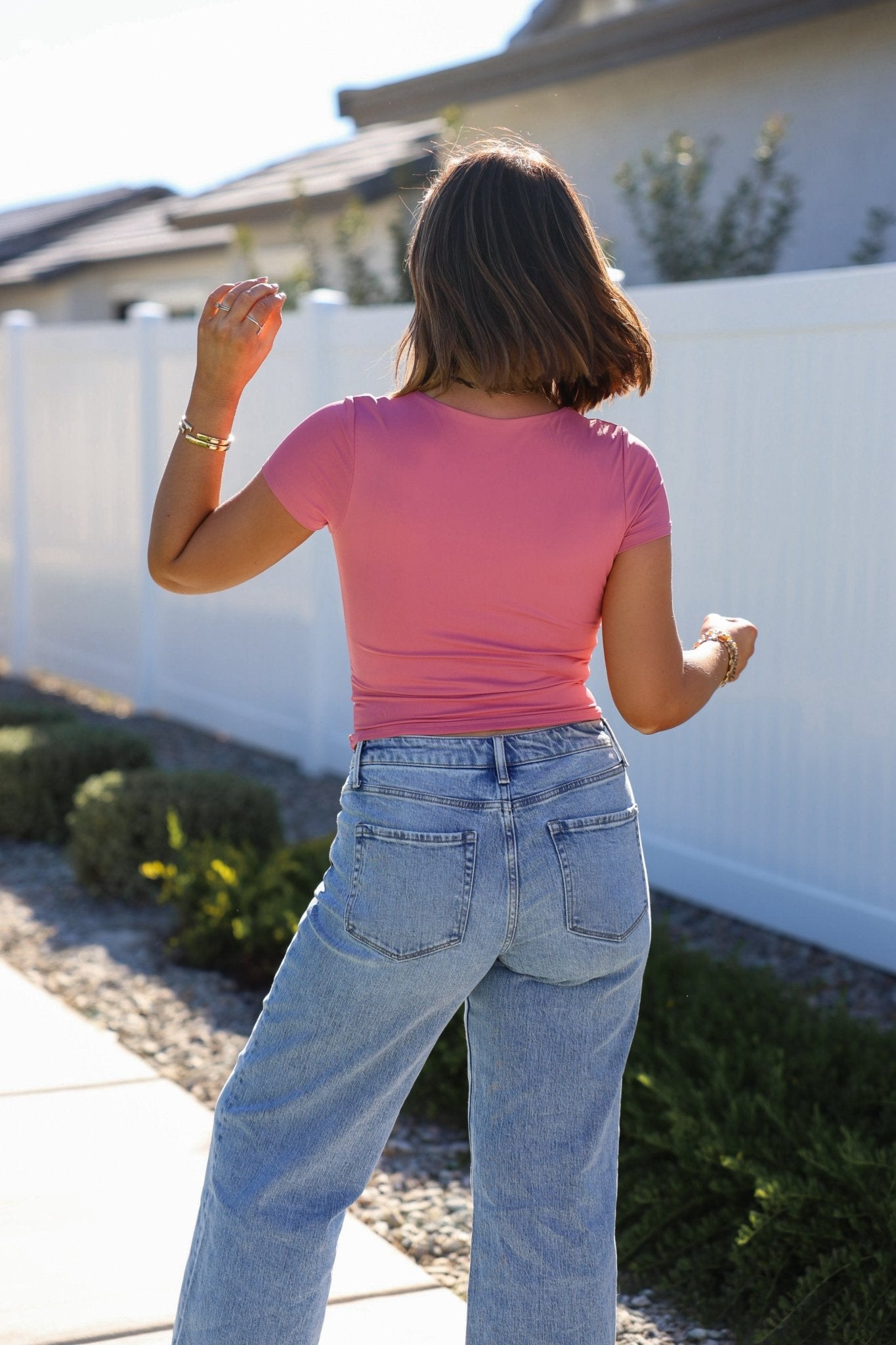 A woman in a Stretchy Pink Double Lined Scoop Neck Top and light blue jeans walks outdoors by a white fence.