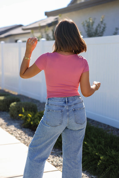 A woman in a Stretchy Pink Double Lined Scoop Neck Top and light blue jeans walks outdoors by a white fence.