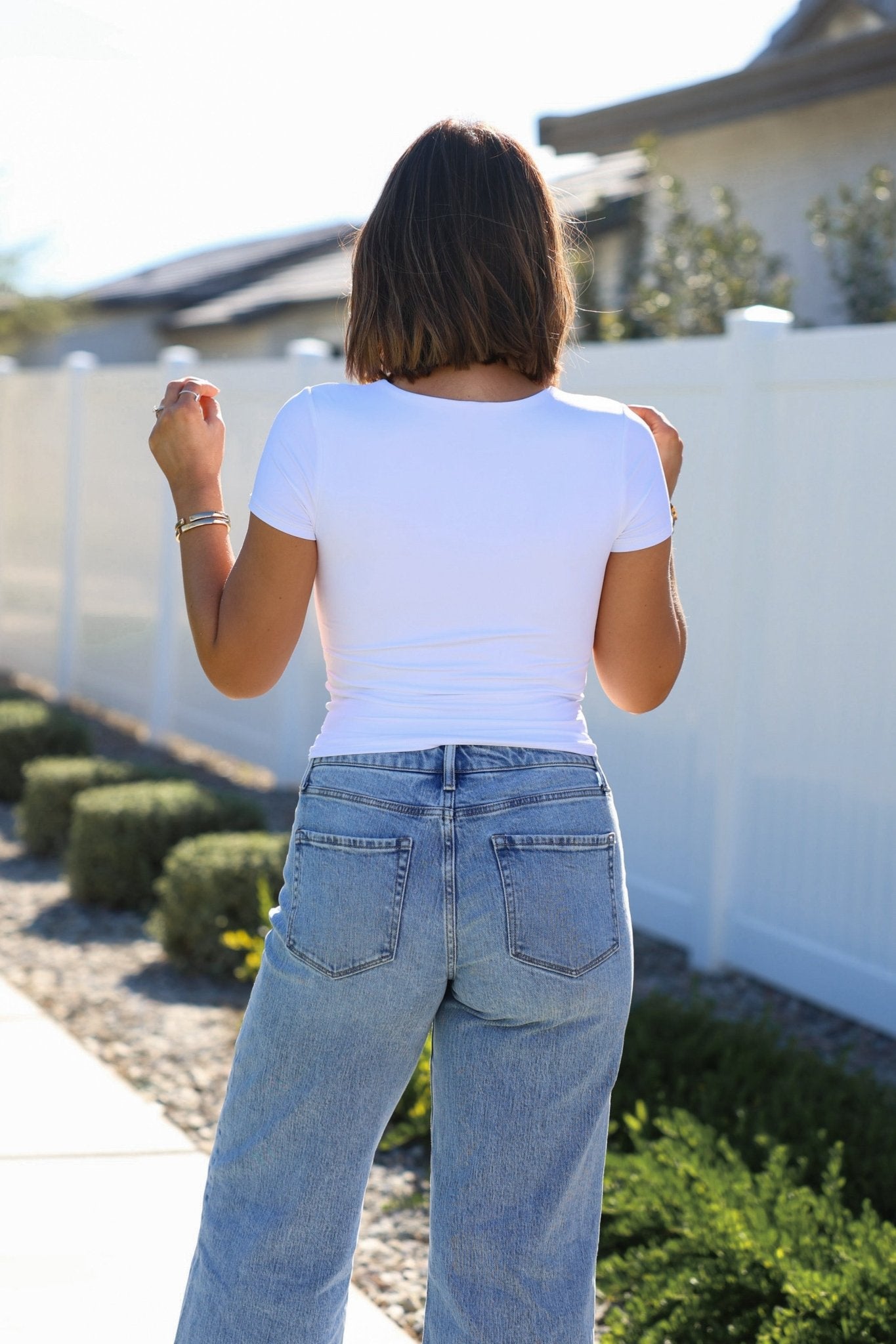 A person with shoulder-length hair stands outside facing away in a Stretchy White Double Lined Scoop Neck Top and blue jeans by a white fence.