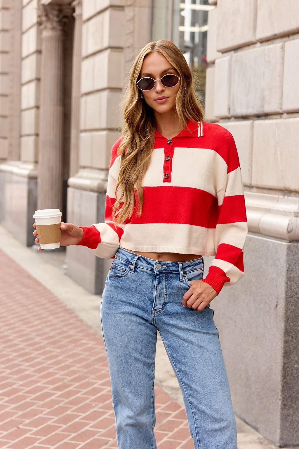 Woman wearing the Striped Button Up Sweater - Red | Pre Order, blue jeans, and sunglasses holds coffee on a city sidewalk.