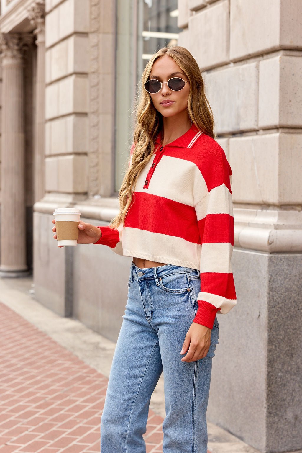 Woman with long hair wears the Striped Button Up Sweater - Red | Pre Order, jeans, and sunglasses while holding coffee on a city sidewalk.