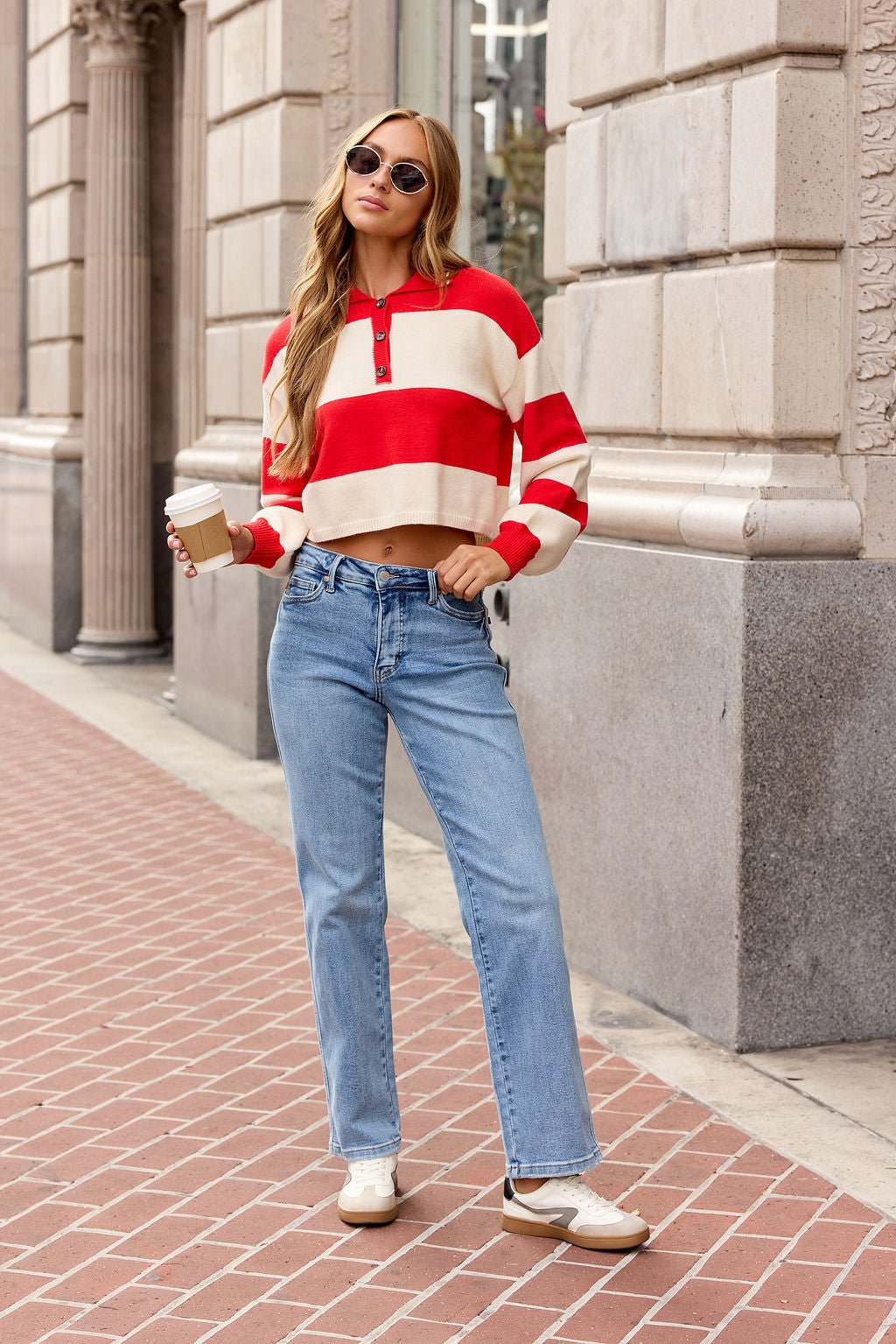 Woman in the Striped Button Up Sweater - Red stands on a city sidewalk with jeans, sunglasses, sneakers, and coffee.