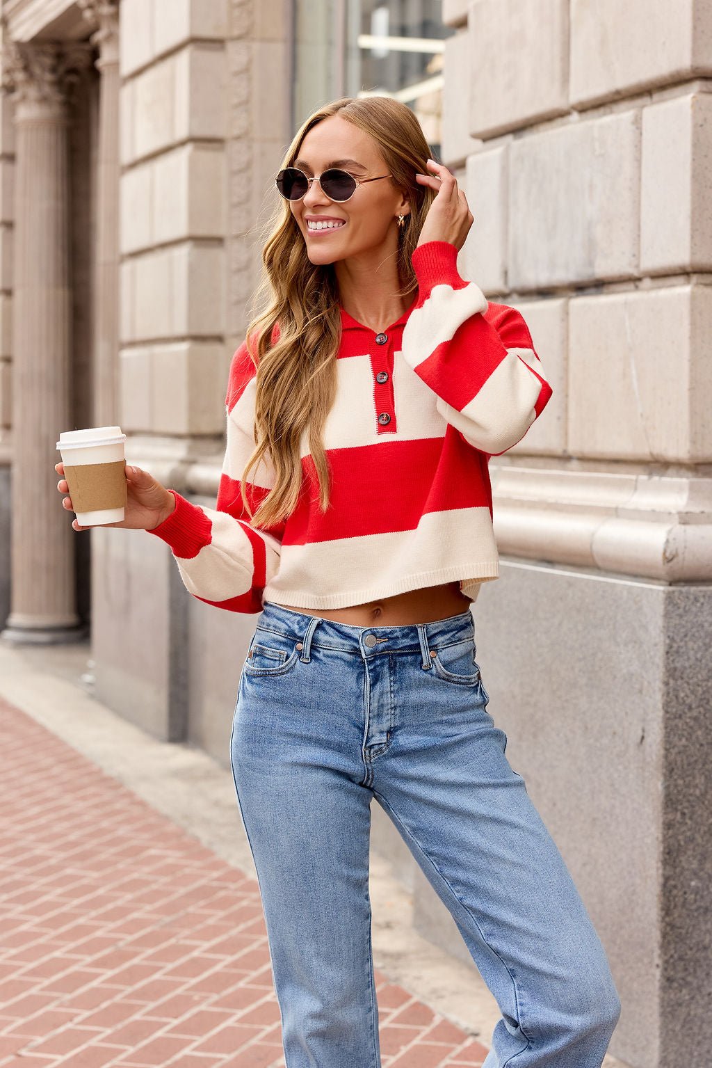 Smiling woman in sunglasses and a Striped Button Up Sweater - Red holds coffee on a city sidewalk.