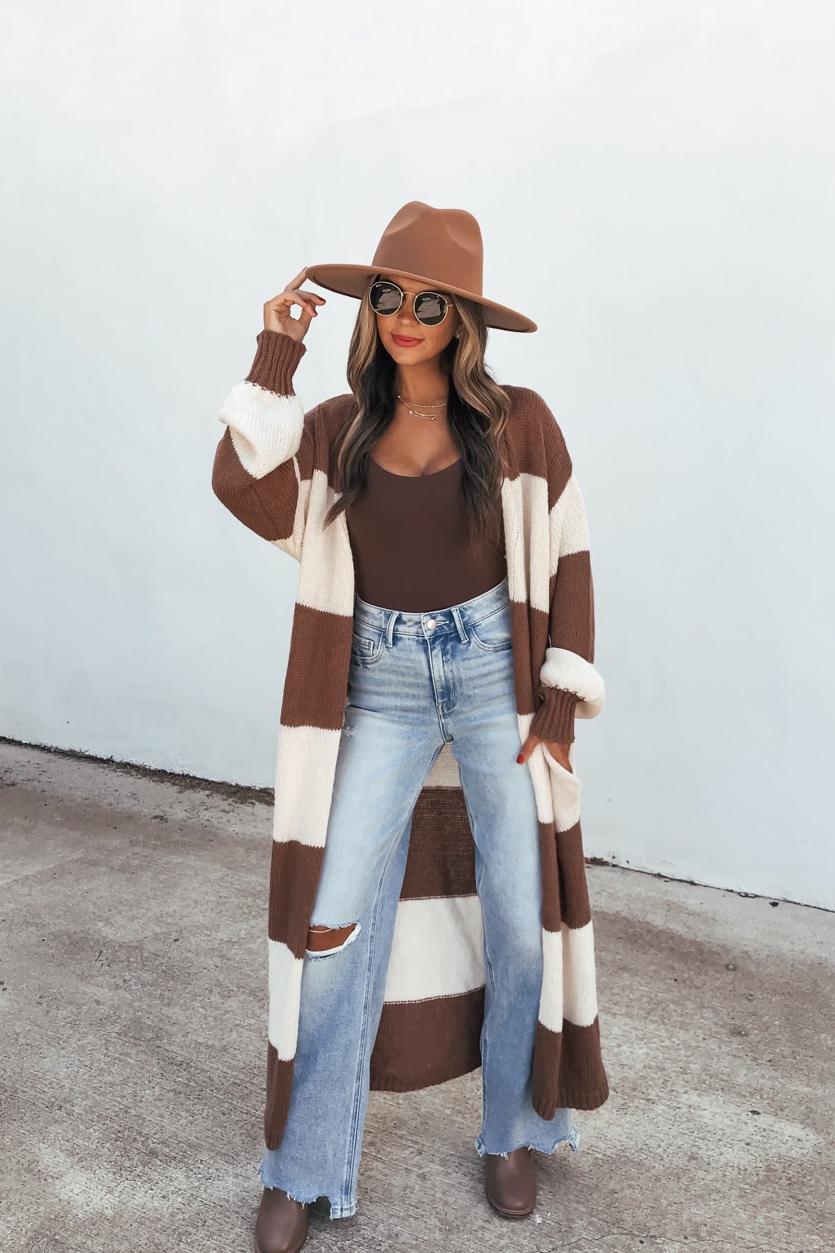 Woman in a brown hat, sunglasses, Striped Duster Cardigan - Brown, brown top, ripped jeans, and boots stands on concrete.