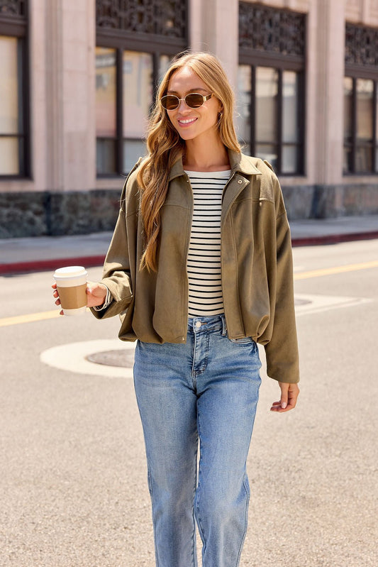 Woman in sunglasses, striped shirt, jeans, and the Suede Bubble Hem Jacket - Olive smiles with coffee on a city street.