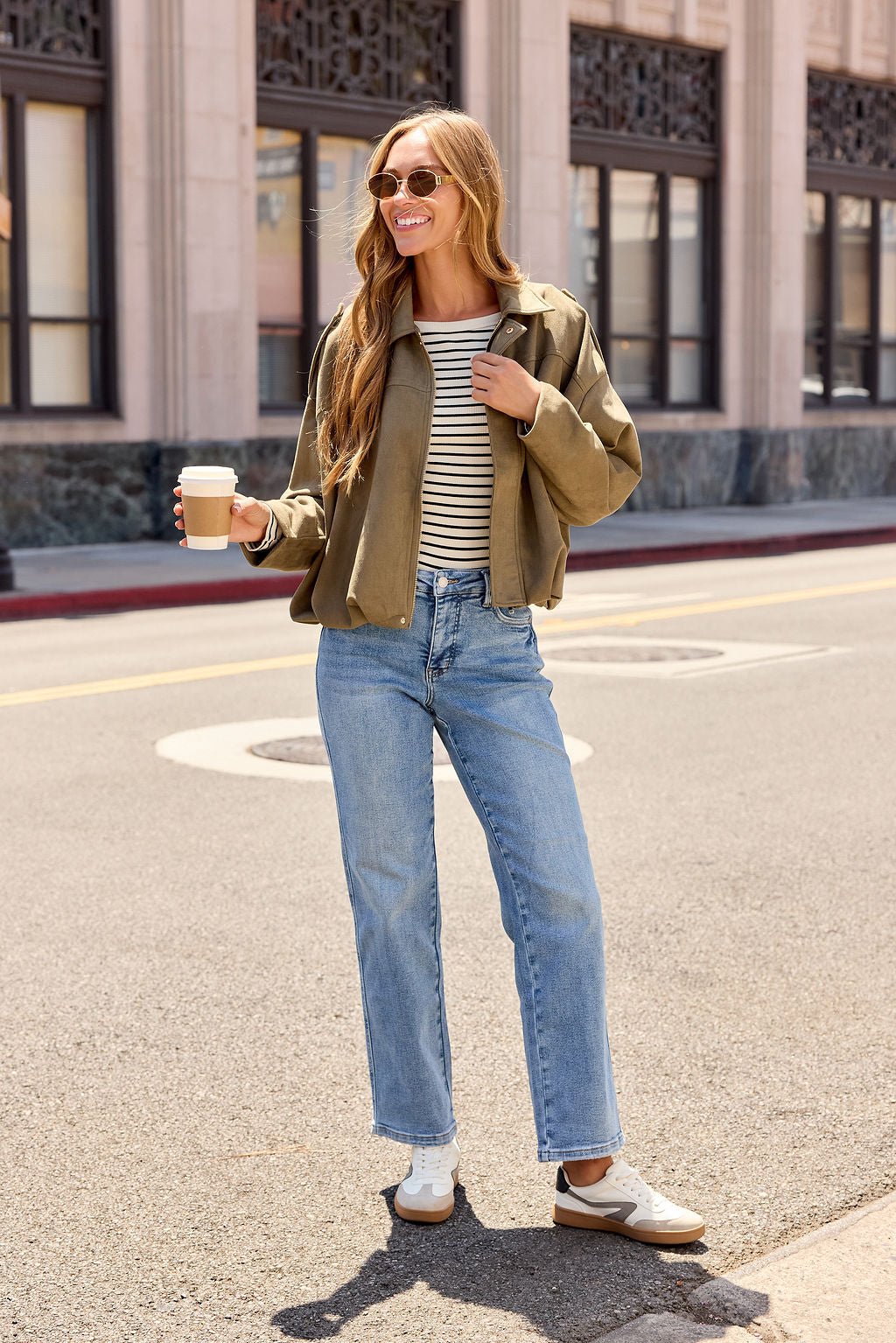 Woman in sunglasses with coffee, layering the Suede Bubble Hem Jacket - Olive over stripes and denim for chic fall city strolls.