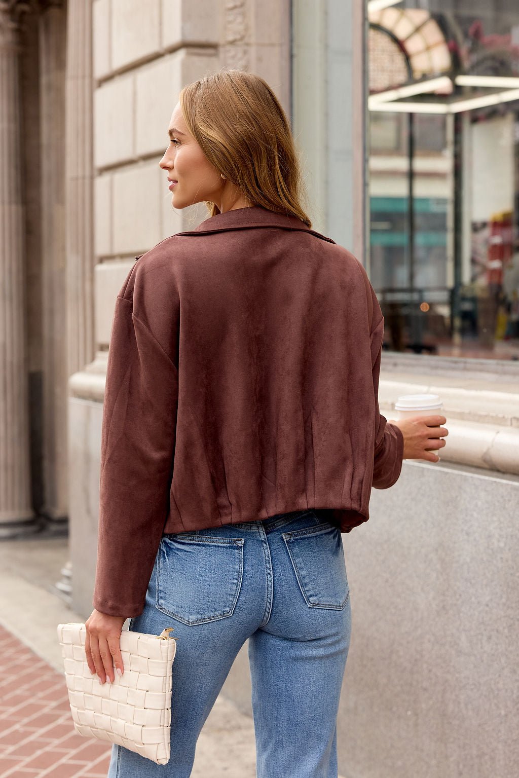 Woman in the Suede Zip Up Jacket - Brown and blue jeans holds a woven clutch and coffee, showcasing this chic layering piece.