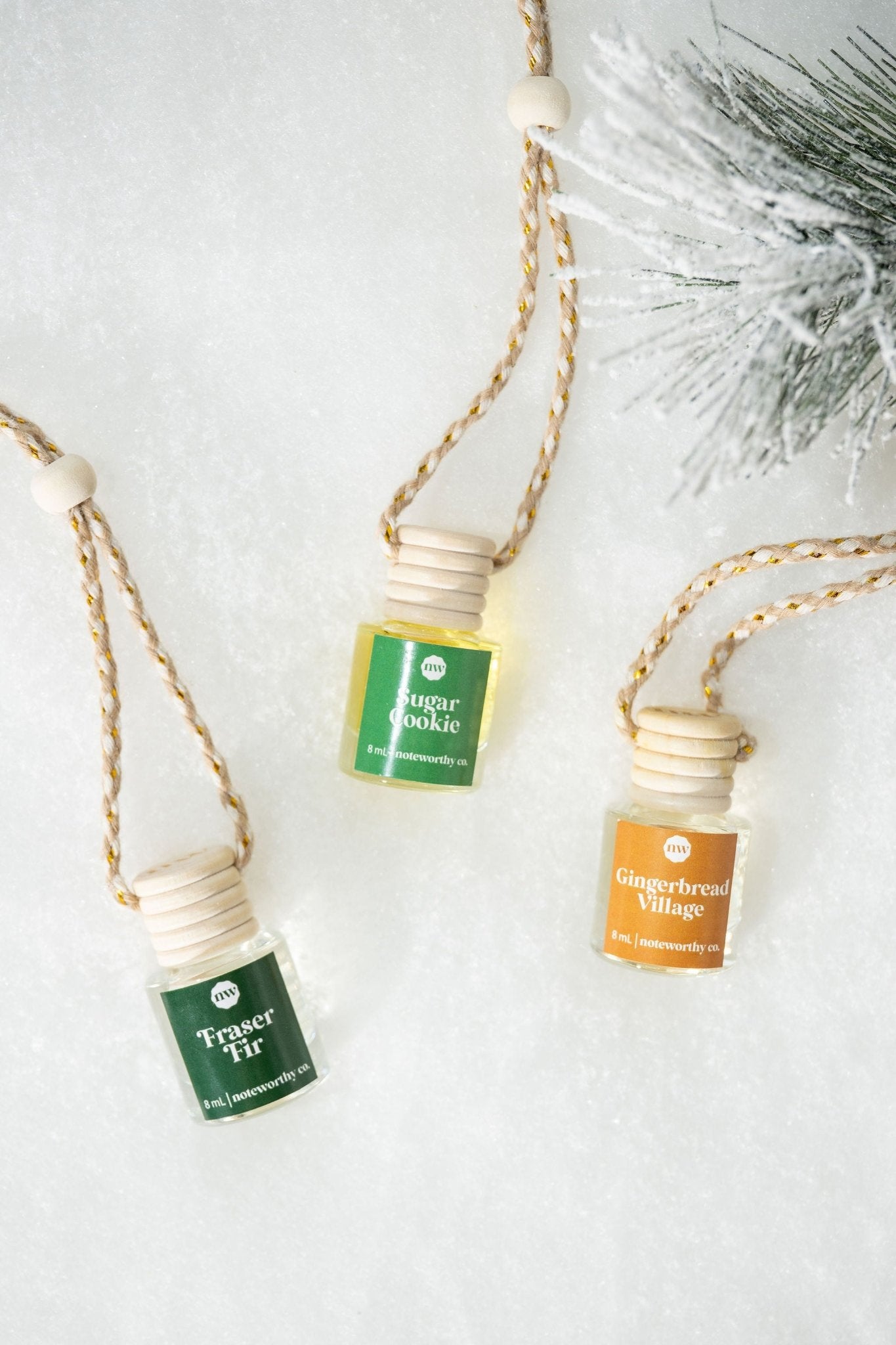 The Sugar Cookie Car Diffuser sits on snow with two other festive mini bottles by a frosted pine branch.