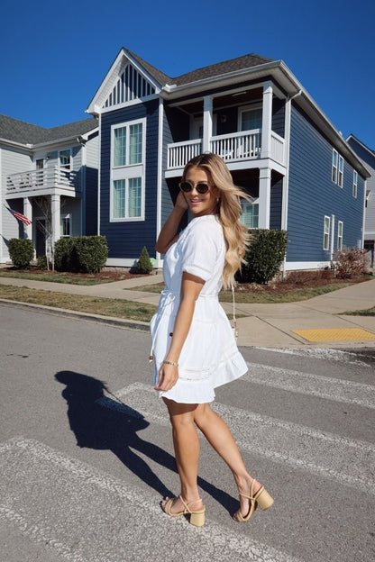 Woman in a Sunday Stroll White Eyelet Mini Dress and sunglasses poses on a crosswalk by blue and white houses.