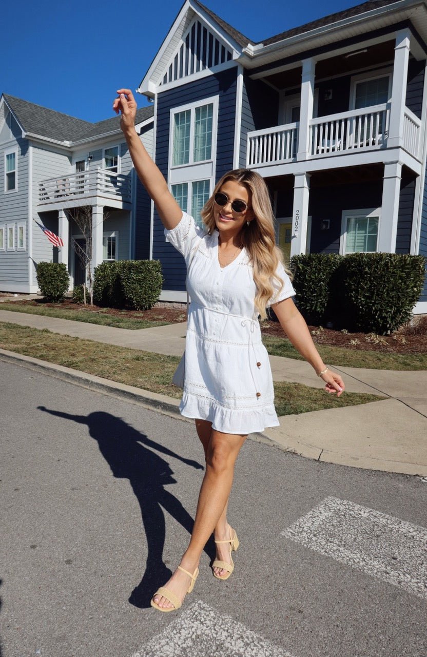 Woman smiles in the Sunday Stroll White Eyelet Mini Dress, posing on a sunny street by blue and white houses.