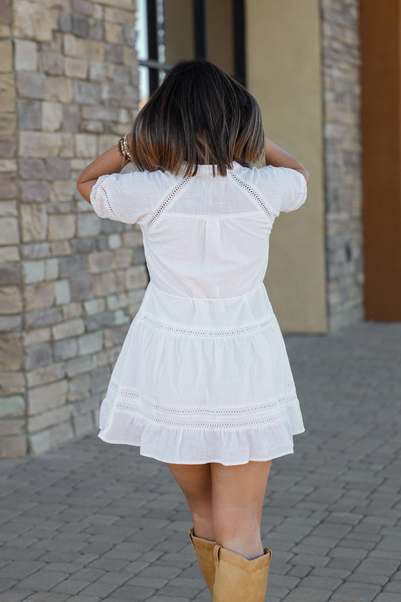 A woman in the Sunday Stroll White Eyelet Mini Dress and tan boots walks on cobblestones, facing away, touching her hair.