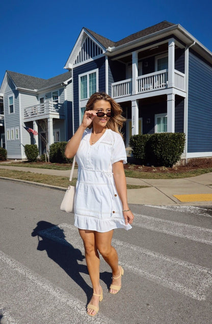 Woman in a Sunday Stroll White Eyelet Mini Dress and sunglasses walks past blue and white houses on a sunny day.
