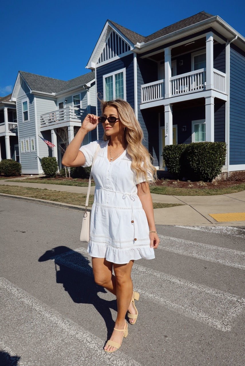 A woman in the Sunday Stroll White Eyelet Mini Dress and sunglasses walks past blue and white houses on a sunny day.