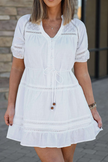 Woman wearing the Sunday Stroll White Eyelet Mini Dress, standing outdoors by a stone wall.