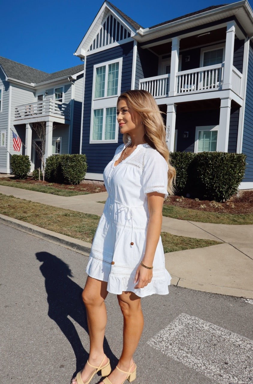 A woman in a Sunday Stroll White Eyelet Mini Dress and sandals walks on a sunny suburban street with blue and white houses.
