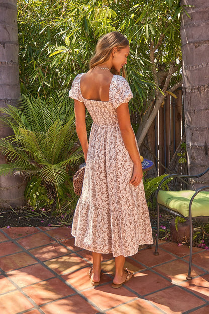 A woman in the Sunlit Grove Floral Midi Dress - Brown stands on a tiled patio, surrounded by tropical plants and palm trees.