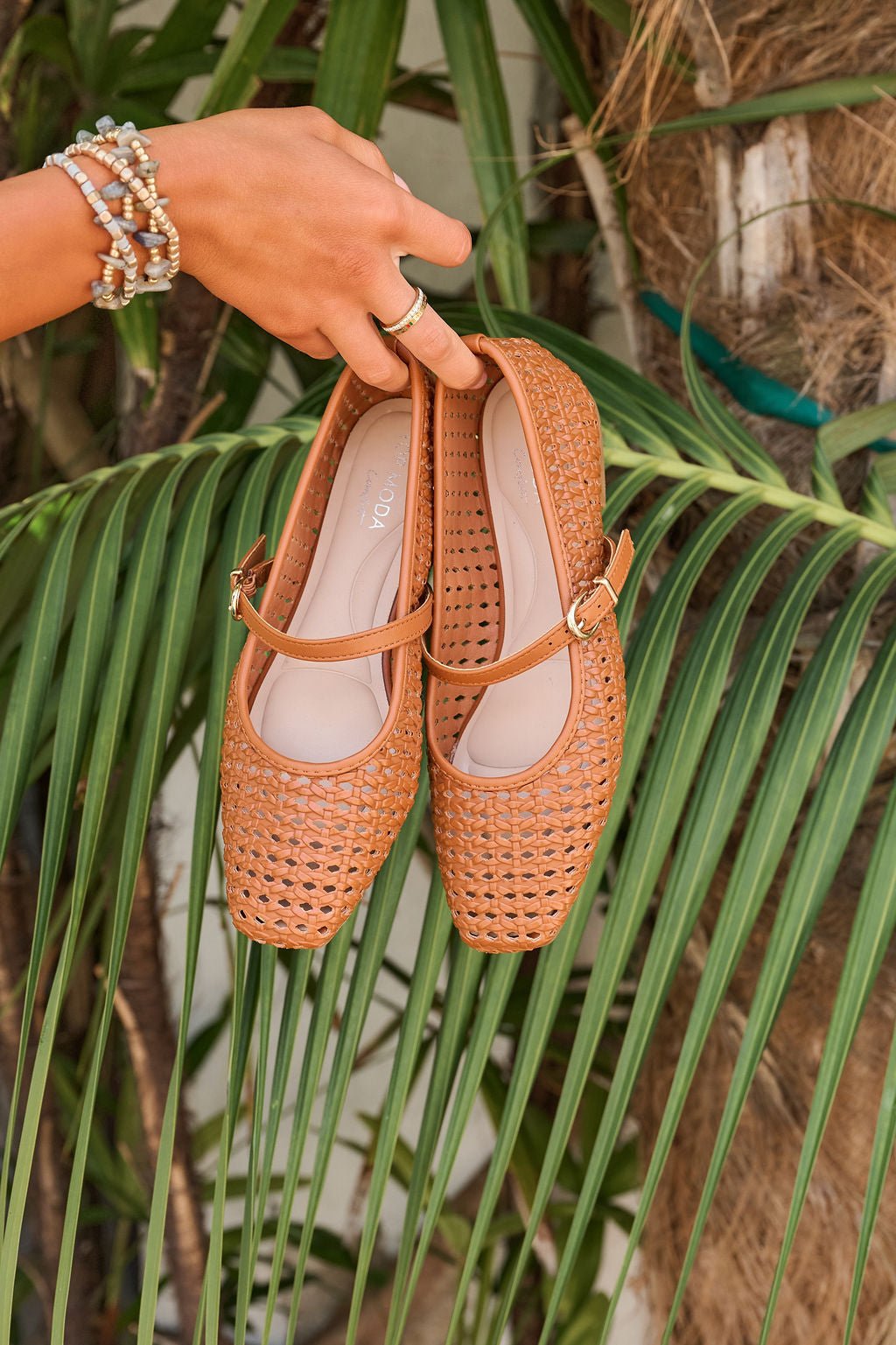 A hand holds Sunny Woven Day Flats - Tan with cushioned insoles above green palm leaves in a tropical outdoor setting.