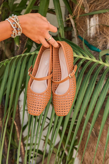 A hand holds Sunny Woven Day Flats - Tan with cushioned insoles above green palm leaves in a tropical outdoor setting.