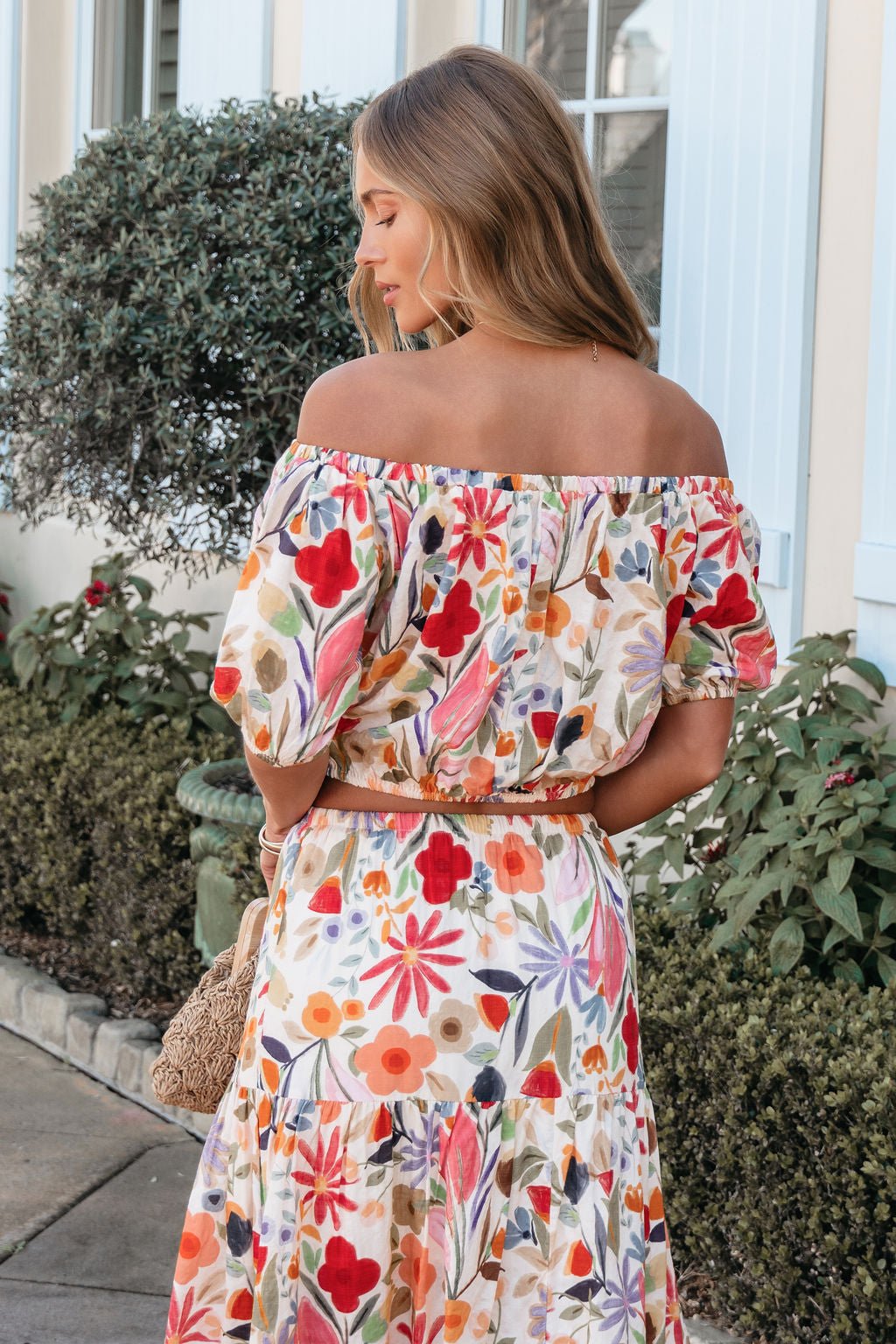 Woman in the Sunset Bloom Multi Print Bubble Hem Top stands outdoors, facing away with wavy hair and a woven bag by her side.