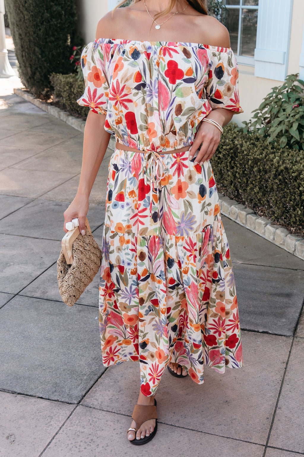 Woman in a Sunset Bloom Multi Print Bubble Hem Top, sandals, and woven handbag standing on a paved walkway.