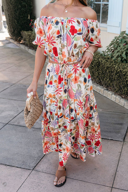Woman in a Sunset Bloom Multi Print Bubble Hem Top, sandals, and woven handbag standing on a paved walkway.