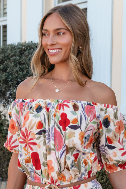 Smiling woman wears the Sunset Bloom Multi Print Bubble Hem Top outdoors near greenery and a white building.