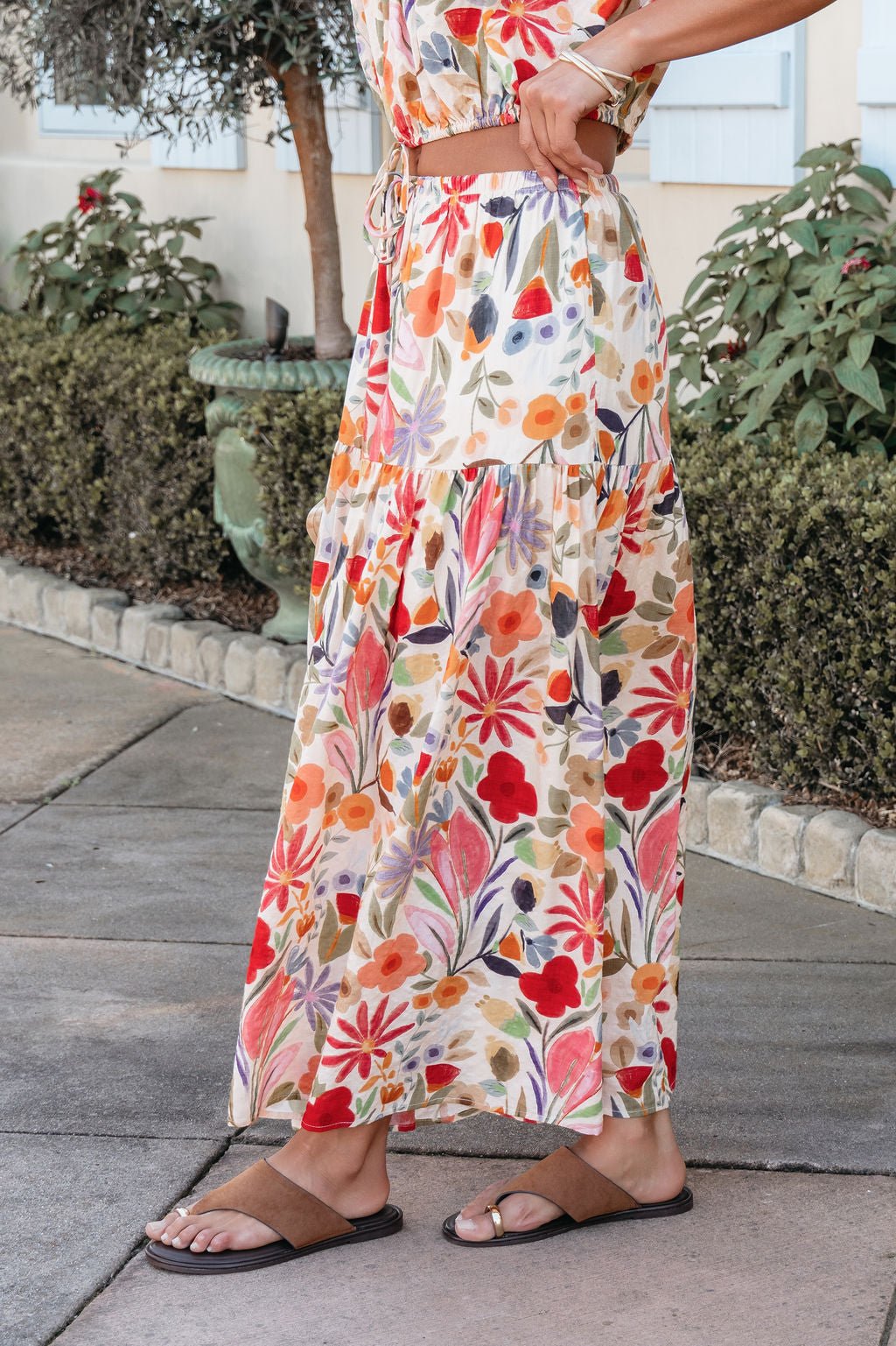 Wearing the Sunset Bloom Multi Print Tiered Maxi Skirt with an elastic waistband and brown sandals near plants on a sidewalk.