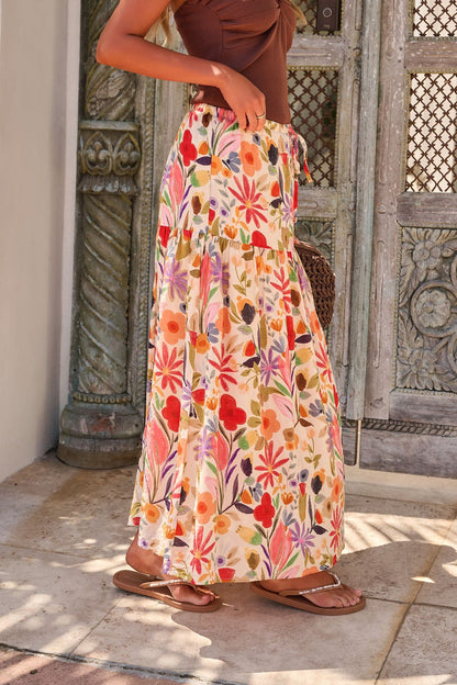 Woman in Sunset Bloom Multi Print Tiered Maxi Skirt with elastic waistband, brown top, and sandals by a carved wooden door.