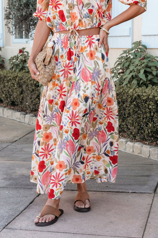 Woman in the Sunset Bloom Multi Print Tiered Maxi Skirt with an elastic waistband, black sandals, and a woven clutch on a sidewalk.