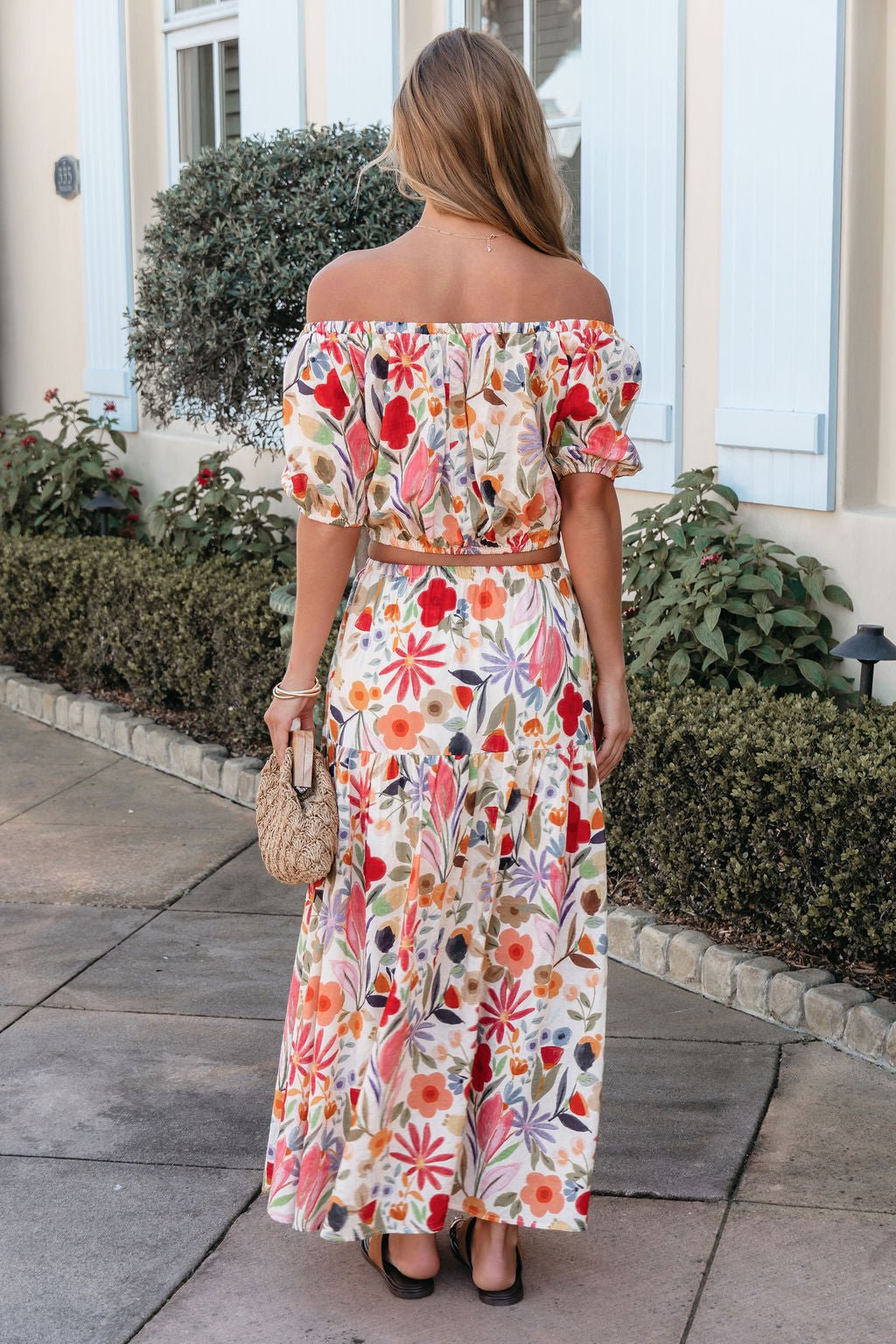 Woman with long hair wears the Sunset Bloom Multi Print Tiered Maxi Skirt, holding a woven bag, standing on a sidewalk facing away.