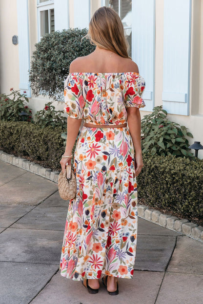 Woman with long hair wears the Sunset Bloom Multi Print Tiered Maxi Skirt, holding a woven bag, standing on a sidewalk facing away.