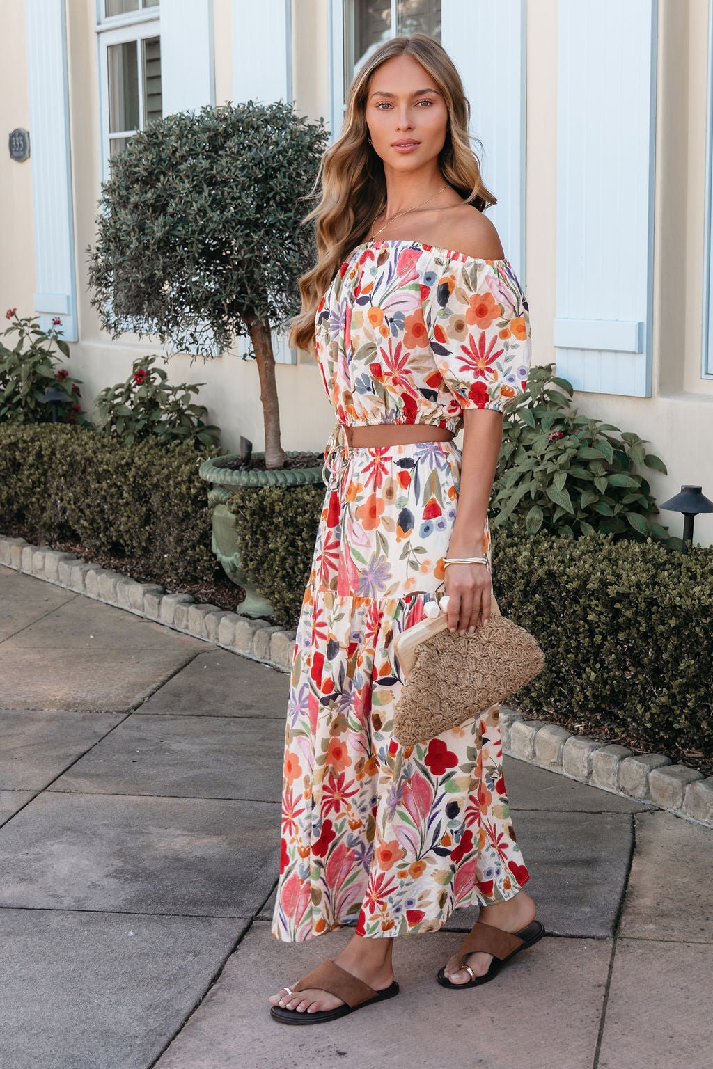Woman in a Sunset Bloom Multi Print Tiered Maxi Skirt with sandals and a woven clutch, standing on a sidewalk near plants.