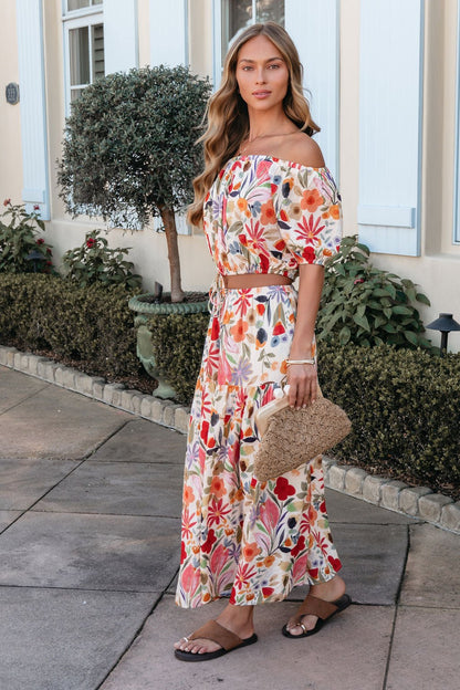 Woman in a Sunset Bloom Multi Print Tiered Maxi Skirt with sandals and a woven clutch, standing on a sidewalk near plants.