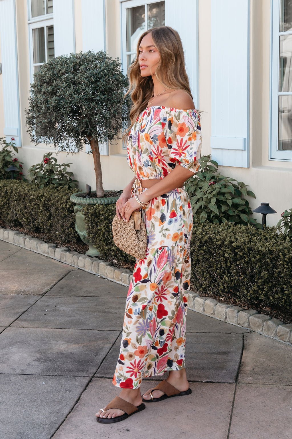 Woman outdoors in the Sunset Bloom Multi Print Tiered Maxi Skirt, holding a woven bag amid greenery and a building.