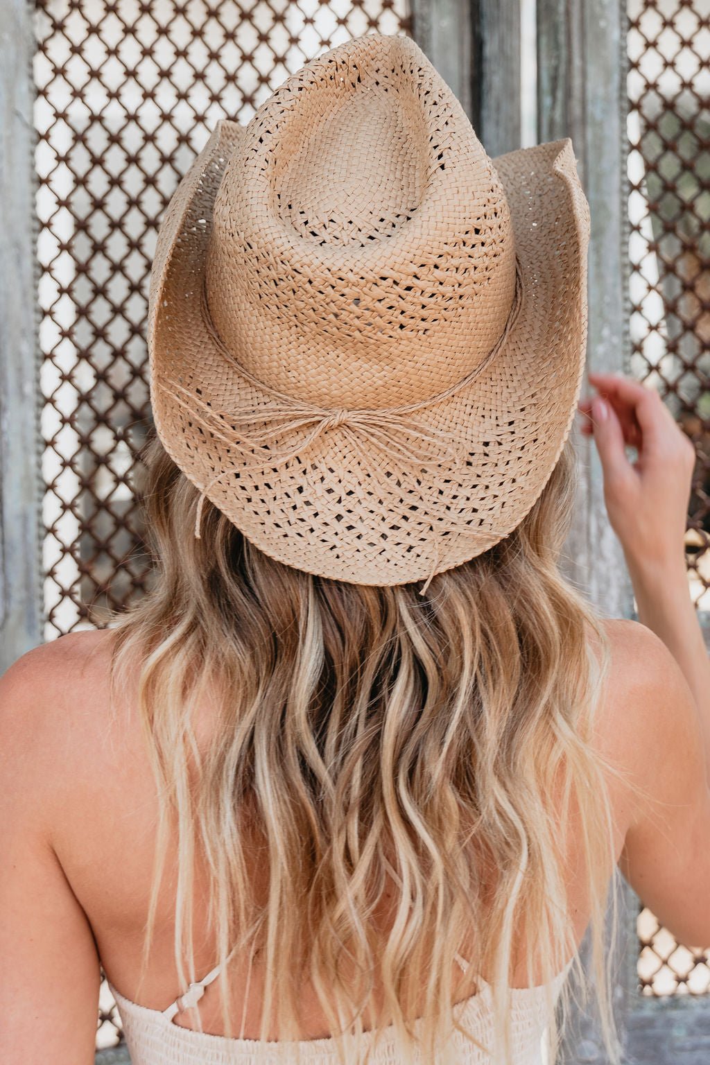 Woman with long wavy blonde hair wears a Sunset Wrangler Straw Cowboy Hat, seen from behind touching a metal mesh gate.