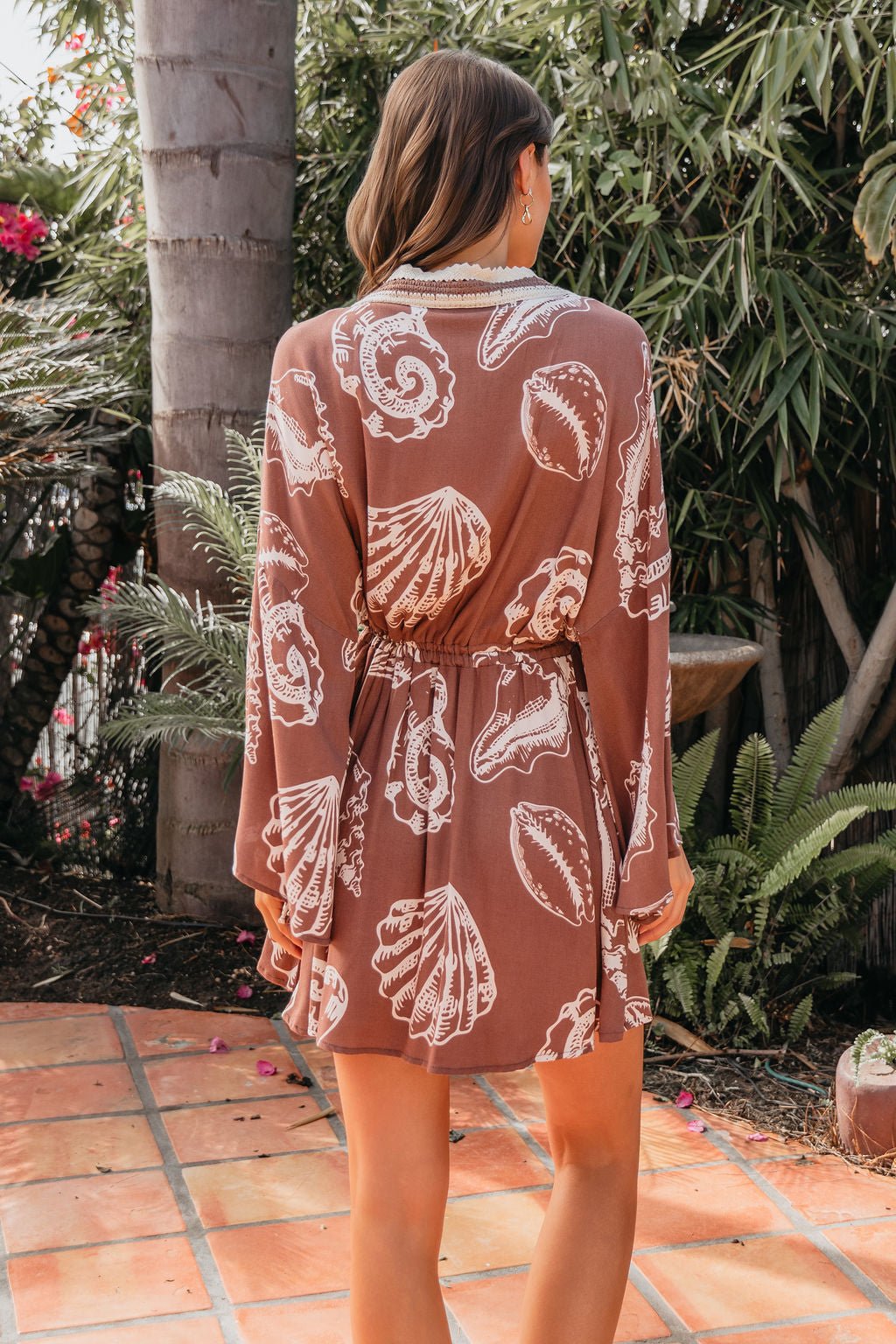 A woman in a Surf Gypsy Crochet Print Dress - Brown stands on a tiled patio outdoors, facing away amid greenery.
