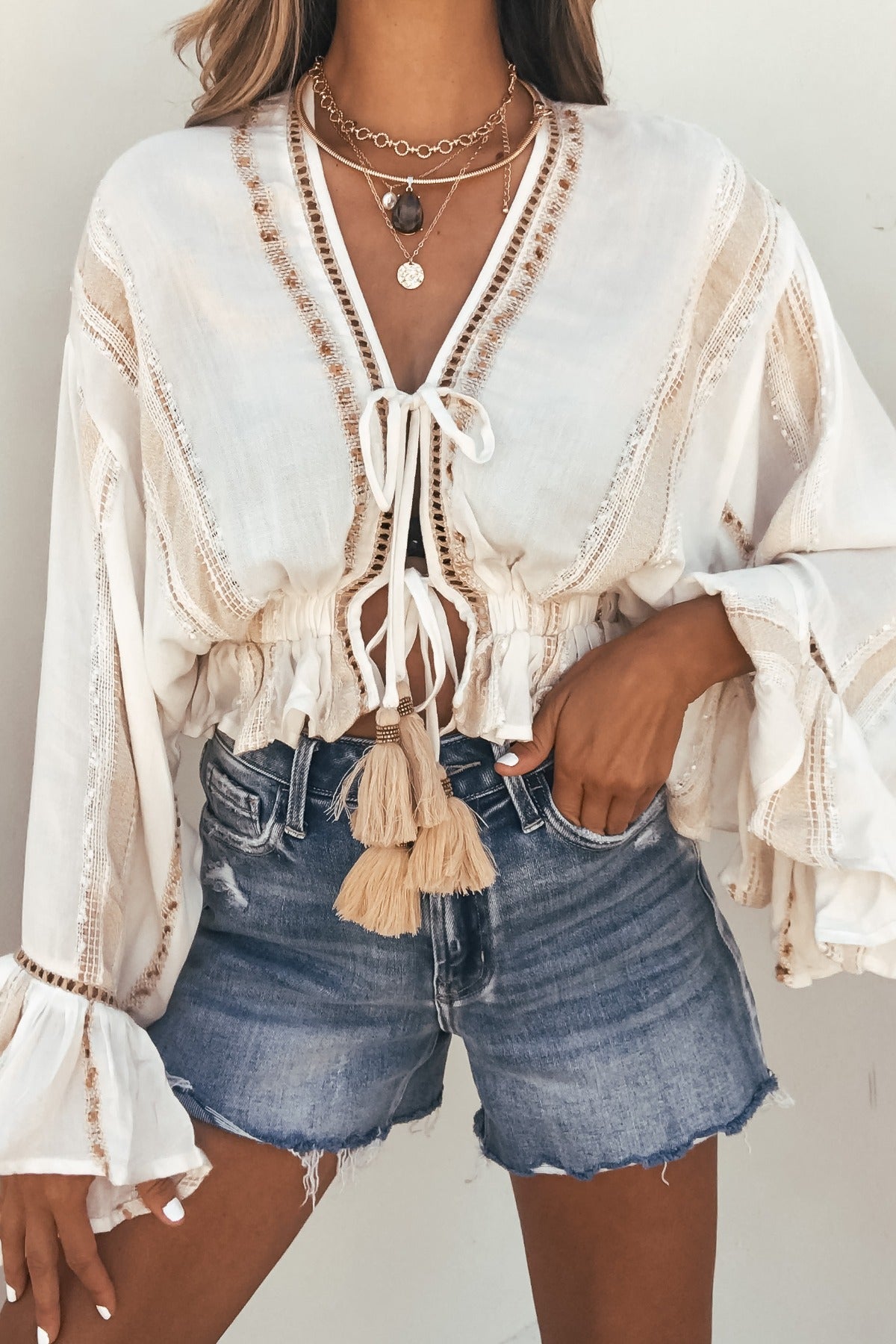 Woman in a Surf Gypsy Gold Dust Stripe Tie Blouse with tassels, layered necklaces, and denim shorts against a plain background.