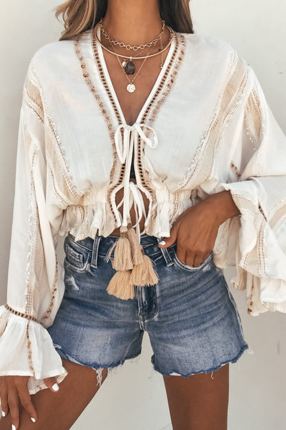 Woman in a Surf Gypsy Gold Dust Stripe Tie Blouse with tassels, layered necklaces, and denim shorts against a plain background.