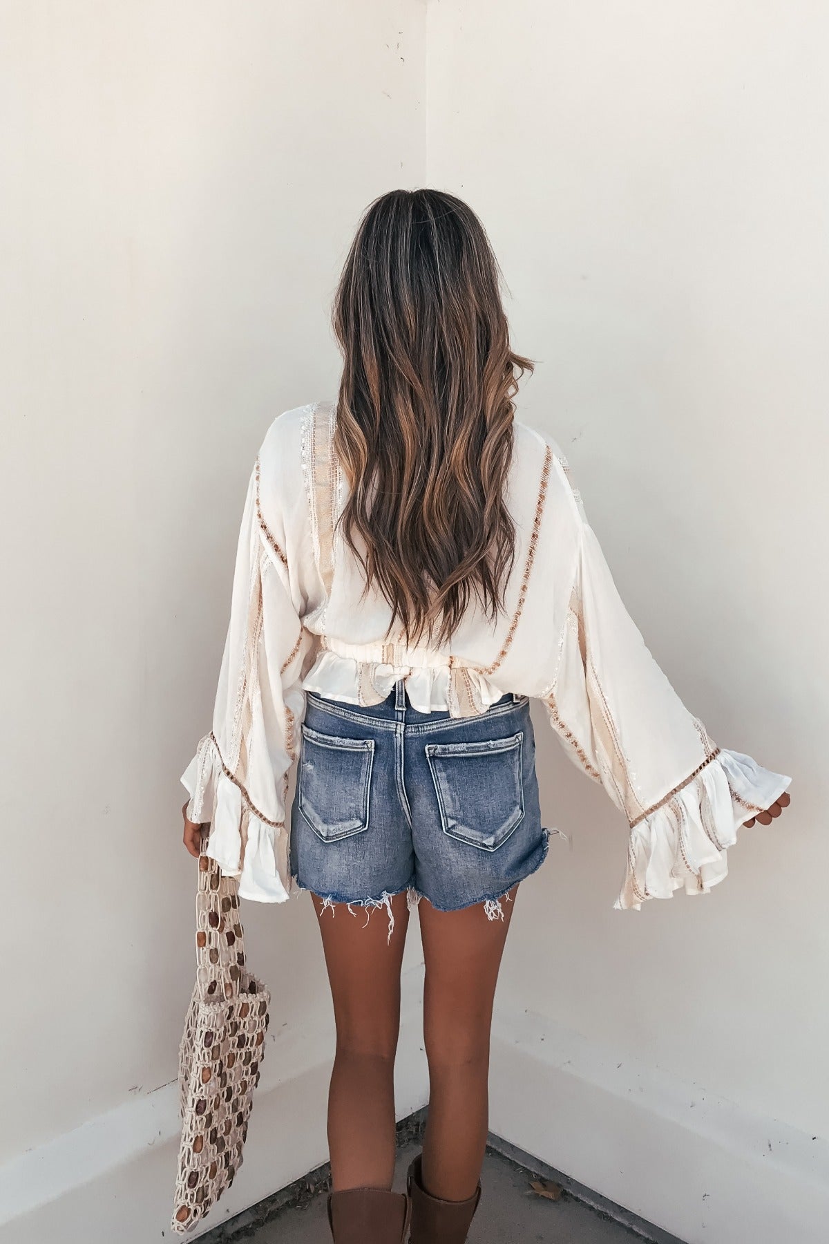 A woman with wavy brown hair in a Surf Gypsy Gold Dust Stripe Tie Blouse and denim shorts stands facing a corner, holding a crochet bag.