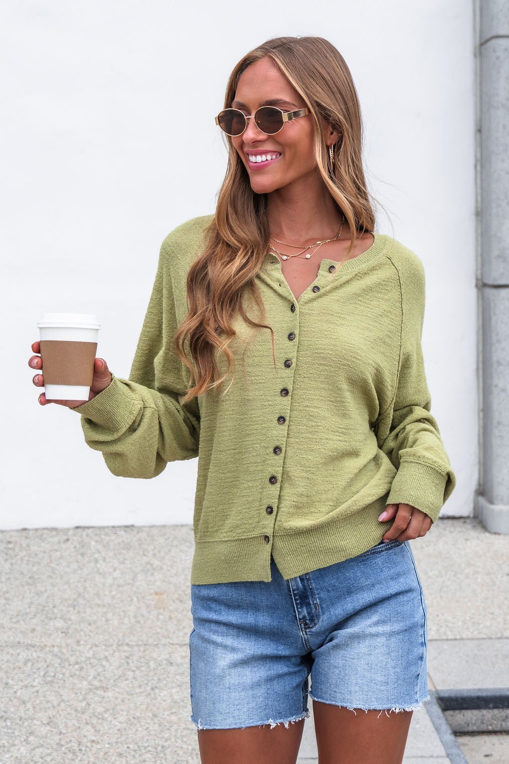 Smiling woman in Sutherlin Button Front Top - Green, denim shorts, and sunglasses holds coffee outdoors against a white wall.