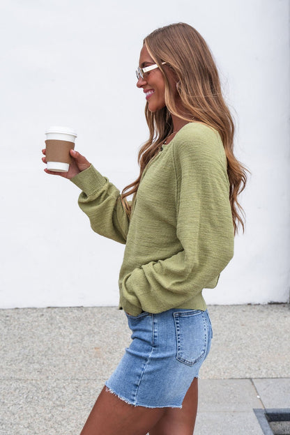 Woman in a green Sutherlin Button Front Top and denim skirt holds a coffee cup, smiling as she walks outside against a white wall.