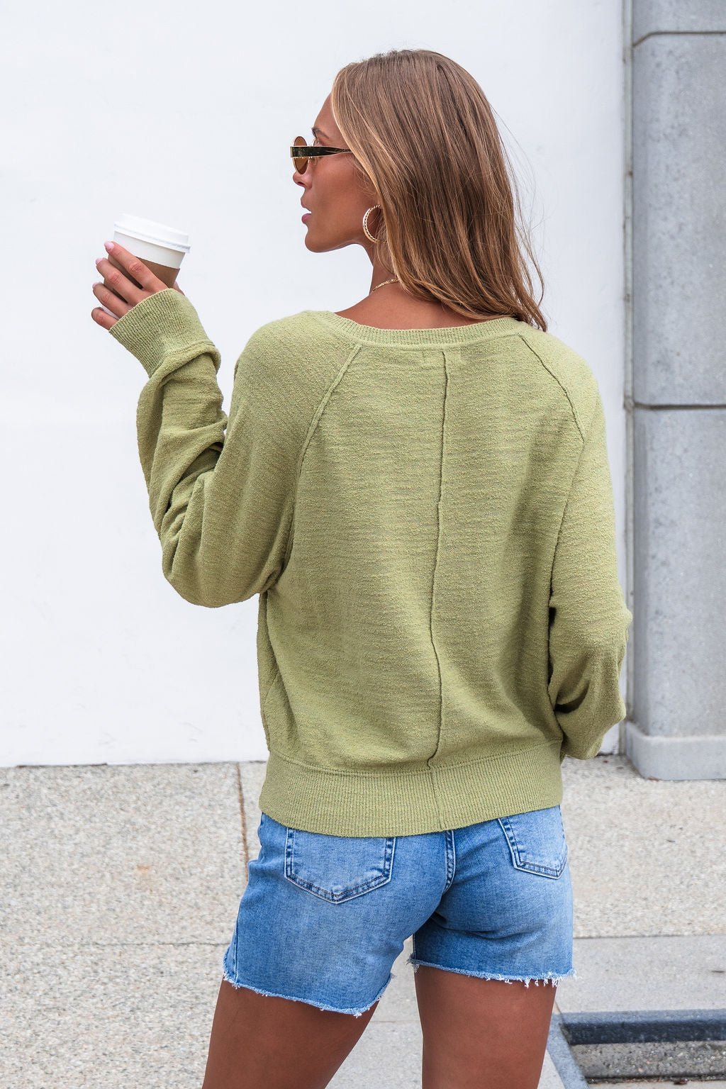 A woman in the Sutherlin Button Front Top - Green and denim shorts holds a coffee, standing outside near a white wall and pillar.
