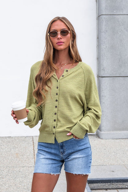 Woman in sunglasses wears the Sutherlin Button Front Top - Green with denim shorts, holding a coffee cup outdoors.