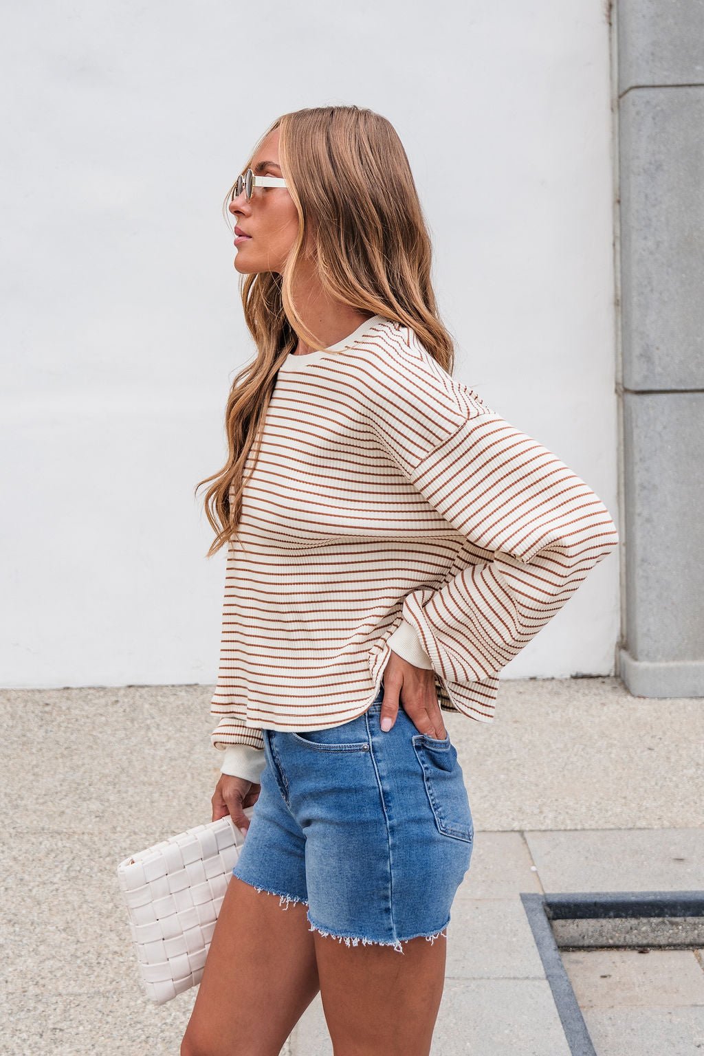 Woman in the Sutton Striped Pullover - Cream with denim shorts stands outside, holding a white clutch, facing left.